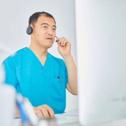doctor-conducting-telemedicine-ime-consultation Male doctor in blue scrubs using a headset while conducting a virtual Independent Medical Evaluation via computer in a well-lit office